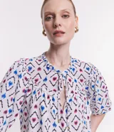 Blusa de chiffon con estampado geométrico en tonos blanco, azul y rosa. Tiene cuello redondo con lazo, mangas cortas abullonadas y volados en el cuello y los puños.