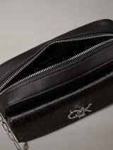 Bandolera negra con monograma CK grabado en toda la superficie. Tiene correa de cadena plateada, cierre superior con cremallera, bolsillo frontal con cierre giratorio y herrajes plateados.