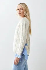 Sweater tejido color crema con diseño de trenzas, corte holgado y camisa celeste a rayas en los laterales.