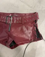 Shorts de efecto cuero color bordó con talle alto. Presenta un cinturón incorporado con hebilla metálica y ojales a lo largo del borde del cinturón y la cintura. Los laterales tienen cordones negros para ajustar el calce.