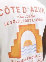 Musculosa oversize de algodón, color blanco, con cuello redondo. Presenta un estampado frontal con temática de la Costa Azul francesa, incluyendo texto en francés e inglés y un gráfico estilizado de un atardecer sobre el mar visto a través de un arco.