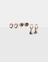 Set de tres pares de aros bañados en oro rosa: un par de aros tipo cuff con strass, un par de aros tipo botón con forma de corazón y cristal color borgoña, y un par de aros colgantes tipo argolla con dije de corazón liso.