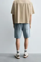 Bermuda de denim de algodón, color gris lavado, con corte relaxed fit y roturas visibles en la parte delantera de ambas piernas. Presenta cinco bolsillos y cierre frontal con cremallera y botón.
