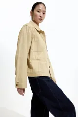 Campera color beige de sarga de algodón, con cuello, botones al frente y canesú en la espalda. Tiene hombros caídos, bolsillos superiores de parche, bolsillos insertados al frente y puños abotonados.
