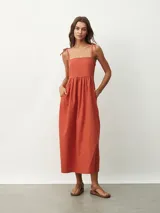 Vestido midi de color naranja, con escote recto y tirantes finos que se anudan sobre los hombros. Posee una cintura entallada con frunces que dan paso a una falda fluida.