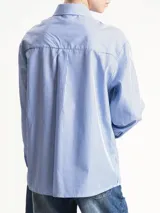 Camisa de manga larga, corte holgado, con cuello camisero y estampado de rayas diplomáticas verticales en blanco y azul. Incluye una corbata ancha integrada en el mismo tejido y patrón, anudada al cuello.