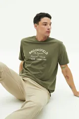 Remera verde militar de manga corta con estampa frontal con el texto "Brookfield The Emporium of Postmodern Activity No 1001 Torino | 1971".