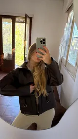 Campera corta de ecocuero marrón oscuro con cierre frontal y dos bolsillos con argollas decorativas.