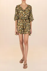 Cover up corto de estilo bohemio con estampado en tonos amarillos, naranjas y negros. Presenta un diseño de tela ligera y traslúcida con escote en V, botones frontales y mangas largas con puños elásticos. Incluye un cinturón de lazo ajustable en la cintura y un ruedo con terminación en volados.