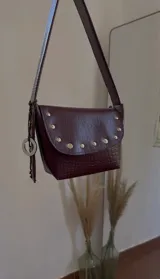 Cartera bandolera color bordó con textura símil cocodrilo, solapa con tachas doradas y correa larga regulable. Incluye charm circular plateado.