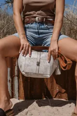 Bandolera color blanco con estampado de manchas grises, correa de cuero marrón y flecos decorativos.