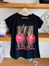 Remera blanca de algodón con cuello redondo y estampado de cerezas rojas con hojas verdes y texto dorado.