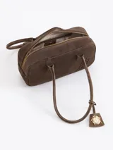 Cartera rectangular de estructura firme confeccionada en cuero descarne vacuno color marrón. Presenta manijas fijas, cierre superior y un charm metálico dorado en forma de vaquita de San Antonio colgando de un tag lateral. Incluye el logo de la marca grabado en relieve en el frente.