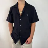 Camisa de manga corta tejida en color negro, con cuello tipo camp y cierre frontal de botones blancos. Presenta un calce holgado y un pequeño logo bordado en el pecho.