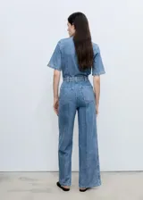 Mono largo de denim azul con cuello camisero, mangas cortas y cierre frontal con cremallera oculta. Incluye cinturón ajustable con hebilla metálica y bolsillo de parche en el pecho.