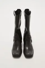 Bota de caña media color negro, estilo motociclista o cowboy, con puntera cuadrada y taco bajo. Presenta una tira ancha con hebillas metálicas alrededor del tobillo.