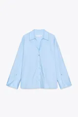 Camisa celeste de popelín con cuello solapa, escote pico y manga larga. Tiene bajo asimétrico con aberturas laterales y cierre frontal con botones.