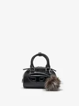 Bolso pequeño tipo bandolera con forma de barril, acabado en cuero negro con textura de piel de lagarto y efecto charol. Presenta el logo 'D' de Diesel en relieve brillante en el frente. Incluye un asa superior rígida y un llavero de pompón de pelo sintético gris y marrón.