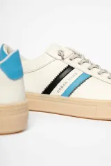 Zapatillas deportivas de cuero sintético color blanco hueso, con suela marrón claro y detalles en azul y negro.