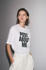 Camiseta blanca de corte amplio, cuello redondo y mangas cortas. Estampado frontal con la frase "Post About Me" en letras negras.
