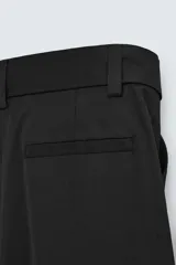 Pantalón negro de corte amplio con cinturón de tela con hebilla metálica. Presenta pliegues frontales, bolsillos laterales y traseros. Cierre frontal con cremallera y botón.
