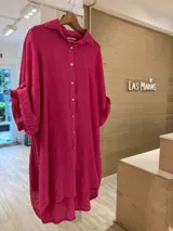 Vestido camisero corto de lino fucsia, fabricado en Italia, con botones en las mangas para recogerlas y botones de nácar. Diseño amplio y cómodo, con un corte irregular que es más largo en la parte trasera que en la delantera.