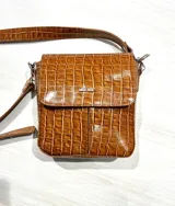 Bandolera marrón con textura simil cuero de cocodrilo, correa larga regulable y logo metálico plateado en el frente.
