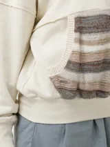 Buzo color beige con cierre frontal parcial y cuello alto. Presenta un bolsillo tipo canguro con diseño tejido a rayas horizontales en tonos marrón, beige y blanco.