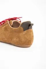 Zapatilla de estilo deportivo color camel, confeccionada en cuero gamuzado. Presenta cordones rojos con detalles blancos y un pequeño refuerzo gris en el talón.