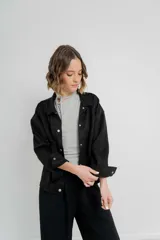 Chaqueta color beige de corte recto, estilo camisa, confeccionada en gamuza con elastano. Presenta cuello camisero, cierre frontal con botones metálicos, bolsillos grandes en el frente y charreteras en los hombros.