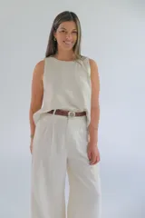 Pantalón de vestir color crudo, de corte palazzo y tiro alto. Presenta pinzas en la cintura y cierre frontal con botón y cremallera. Incluye cinturón trenzado marrón con hebilla redonda plateada.