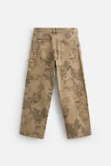 Pantalón de jean color beige con estampado abstracto floral en tonos marrones, de corte amplio, con bolsillos delanteros y traseros, y detalle de tira multifunción en la pierna. Cierre frontal con cremallera y botón.