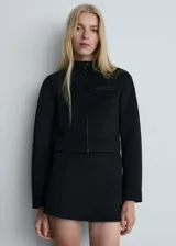 Chaqueta corta negra con cuello mao, cierre frontal con cremallera, dos bolsillos laterales y un bolsillo de ribete en el pecho.