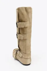 Bota alta color beige de serraje con puntera redondeada. Presenta tres tiras con hebillas metálicas en la parte delantera y caña.