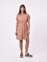 Vestido camisero corto color tostado, con cuello camisero, mangas cortas con dobladillo, cierre frontal con botones, cinturón de lazo y bolsillos en el pecho.