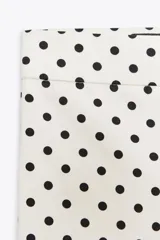 Pantalón largo de tiro alto con estampado de lunares negros sobre fondo blanco. Tiene falsos bolsillos de vivo en la espalda, detalle de solapa en el delantero y bajo acampanado. Se cierra con cremallera y gancho metálico.