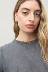 Blusa azul grisáceo de algodón con bordados frontales y terminaciones festoneadas. Tiene mangas cortas con frunces y cuello redondo con detalle de puntilla.