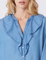 Blusa de lyocell color azul claro, con escote en V y volados en el cuello y la parte frontal. Tiene mangas largas con puños elásticos y cierre de botones en la parte delantera.