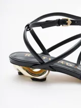 Sandalias negras de cuero con tiras finas, taco geométrico dorado y pulsera al tobillo.