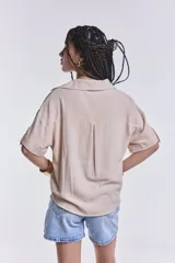 Camisa color beige de corte cropped, con cuello clásico, cierre frontal con botones y dos bolsillos de parche en el pecho.