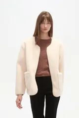 Chaqueta corta de tejido bouclé o pelo sintético color crema, con cuello redondo, cierre frontal metálico y dos bolsillos frontales amplios.