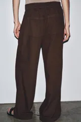 Pantalón marrón de tiro medio, corte ancho y largo cropped. Confeccionado en algodón, con cintura con trabillas y elástico en la espalda, bolsillos laterales y traseros, pinzas en el delantero y cierre frontal con cremallera, botón y ganchos metálicos.