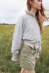 Pollera corta de denim color verde oliva, estilo cinco bolsillos, con cierre y botón frontal. Presenta apliques de tachas metálicas doradas y plateadas en forma de estrella o brújula distribuidas en el frente.