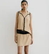 Blusa color beige con cuello camisero y botones frontales, sin mangas y con ribete negro en cuello, sisas y ruedo.