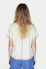 Remera beige de corte holgado con cuello redondo y mangas cortas. Presenta un pequeño estampado de un sol azul en el centro del pecho y costuras en color rojo en cuello, mangas y ruedo.