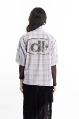 Camisa unisex de manga corta con estampado a cuadros en tonos verde, negro, blanco y morado. Presenta un corte boxy y un estampado en serigrafía en el pecho.
