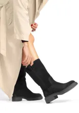 Bota alta de cuero negro, estilo caña media/larga, con caña ancha y suela track gruesa de goma negra. Presenta un diseño liso y minimalista, con una pequeña tira de tela con texto en el borde superior de la caña.