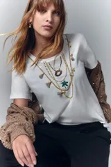 Remera blanca de algodón con cuello redondo y mangas cortas. Presenta un estampado en el escote simulando collares de estilo boho, con bordados y apliques de lentejuelas y flecos.