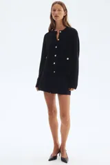 Cárdigan corto negro de punto acanalado, con cuello redondo, manga larga y botones metálicos dorados en el frente. Posee bolsillos de parche con botón en la parte inferior delantera.