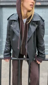 Chaqueta de cuero ovino color marrón, con cuello clásico, cierre asimétrico con cremallera, solapas con botones a presión, múltiples bolsillos con cremallera y cinturón ajustable en la cintura.